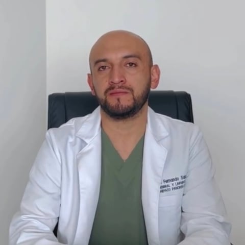 Male healthcare professional in white lab coat and green scrubs seated in chair, wearing stethoscope around neck