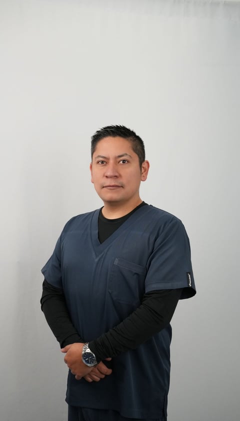 Man in dark blue medical scrubs and black long-sleeved shirt standing against white background, wearing a watch and looking at camera
