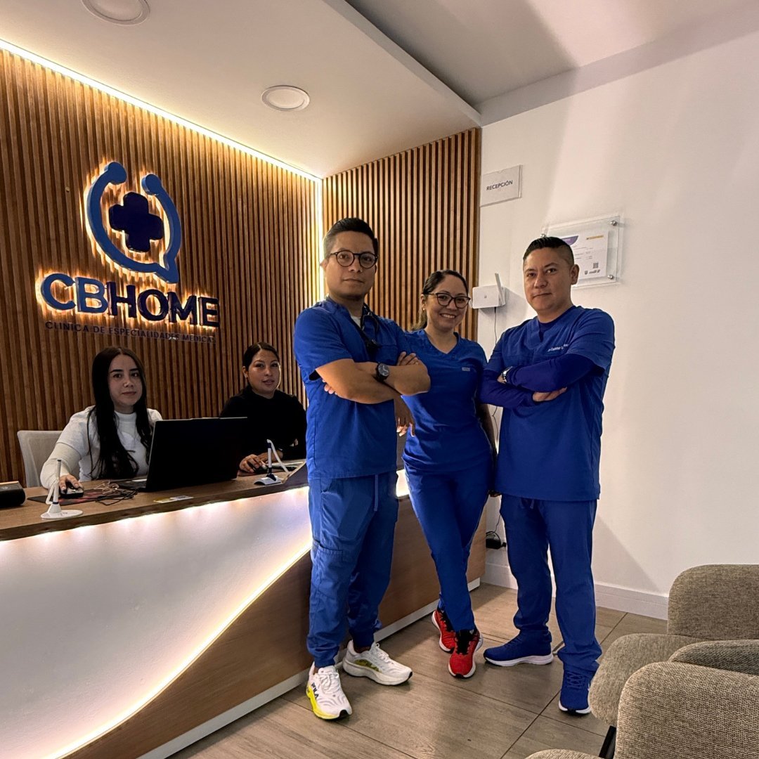Three men in blue medical scrubs pose together in a modern clinic reception area with GB Home logo on wood-paneled wall